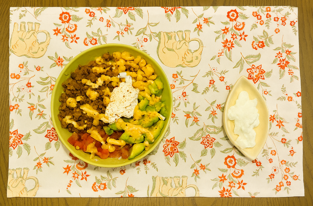 A colourful burrito bowl with avocados, tomatoes, minced lamb and sweetcorn on top of a base of seasoned cilantro rice. Served with a side of sour cream.